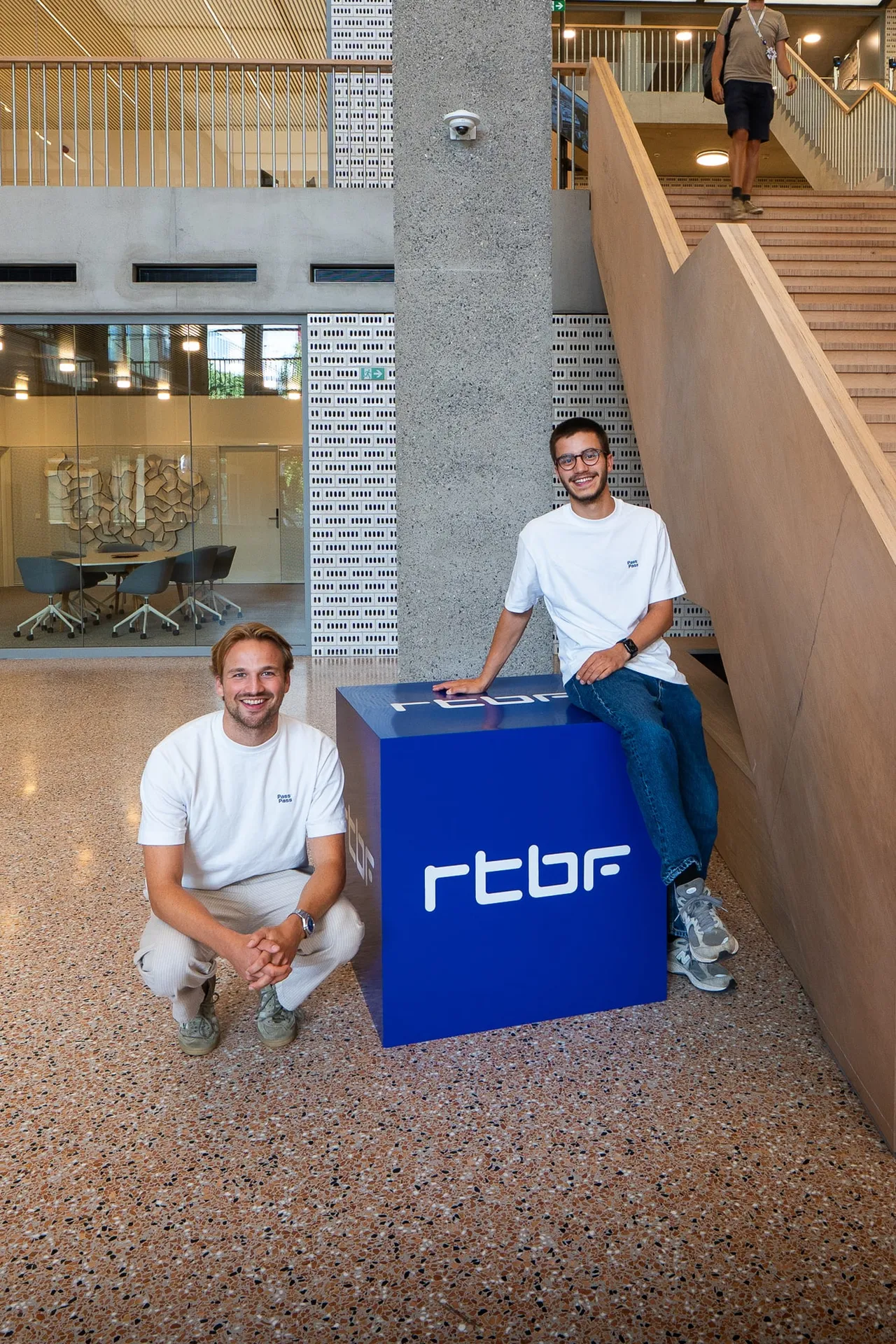 Deux personnes posant à côté d’un cube aux couleurs de la RTBF, dans le hall d’un bâtiment.