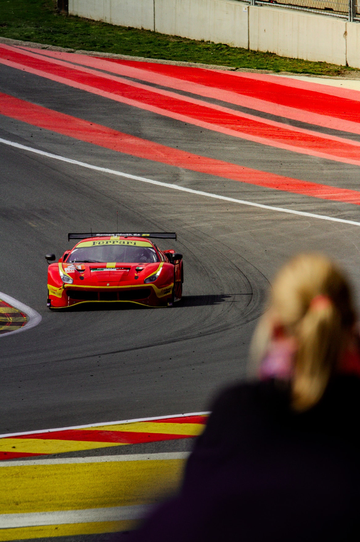 Une Ferrari 488 GT3 rouge et jaune à la corde d’un virage, vue par-dessus l’épaule d’une spectatrice.