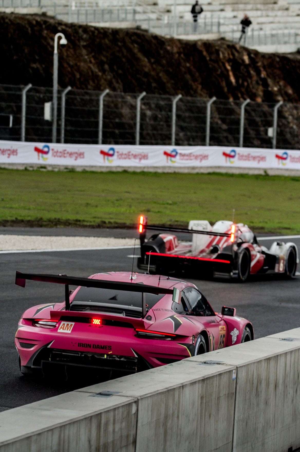 La Porsche 911 GT3 R rose n° 85 des Iron Dames vue de trois quarts arrière, doublée par une Hypercar rouge et blanche.