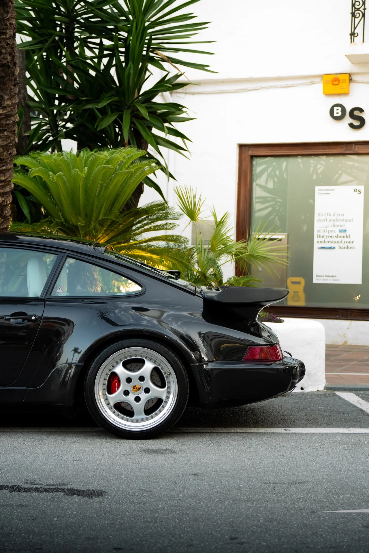 Trois quarts arrière d’une Porsche 964 Turbo noire, aileron déployé et jante Cup polie au premier plan.