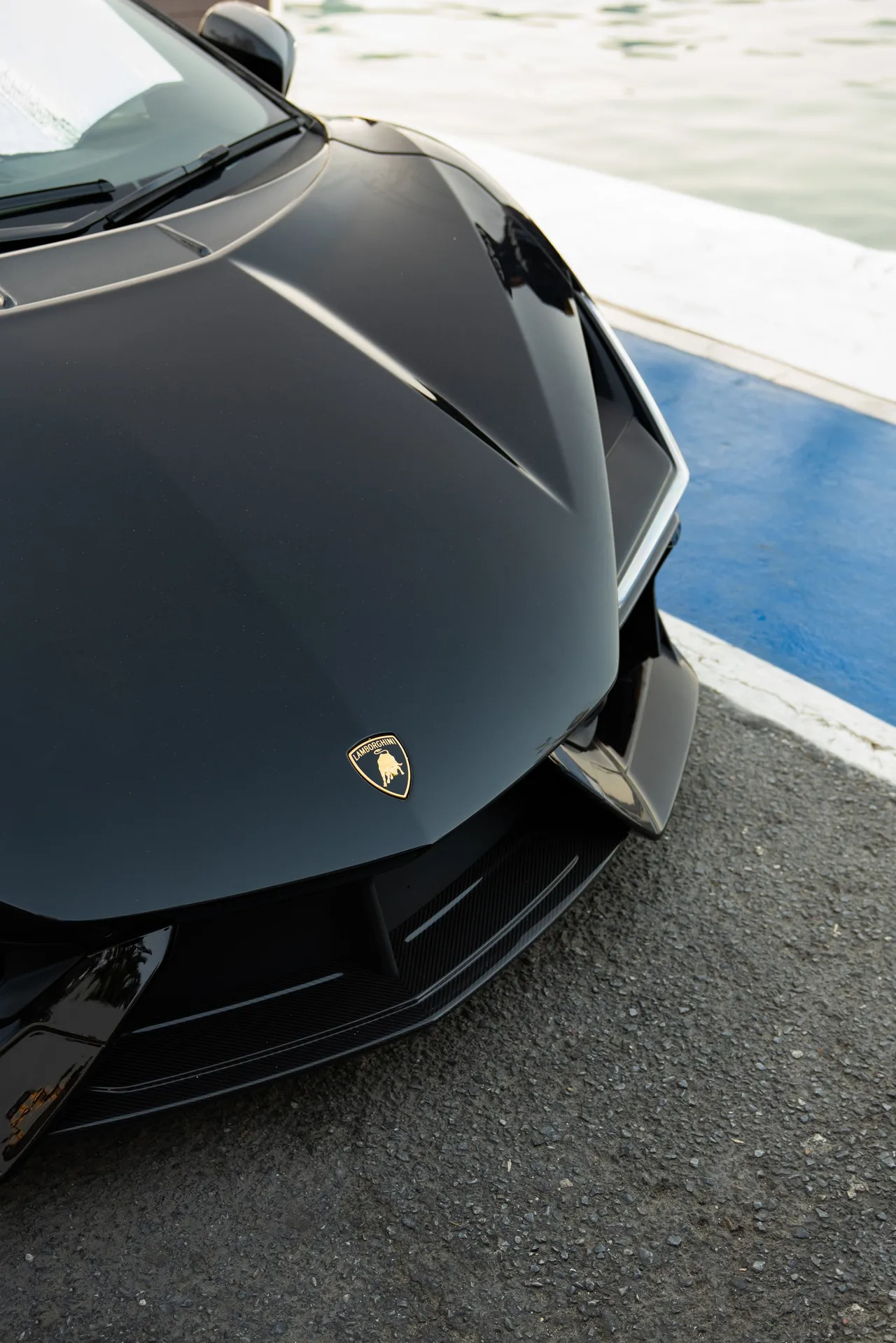 Capot avant d’une Lamborghini noire au bord d’un quai, l’eau du port en arrière-plan.