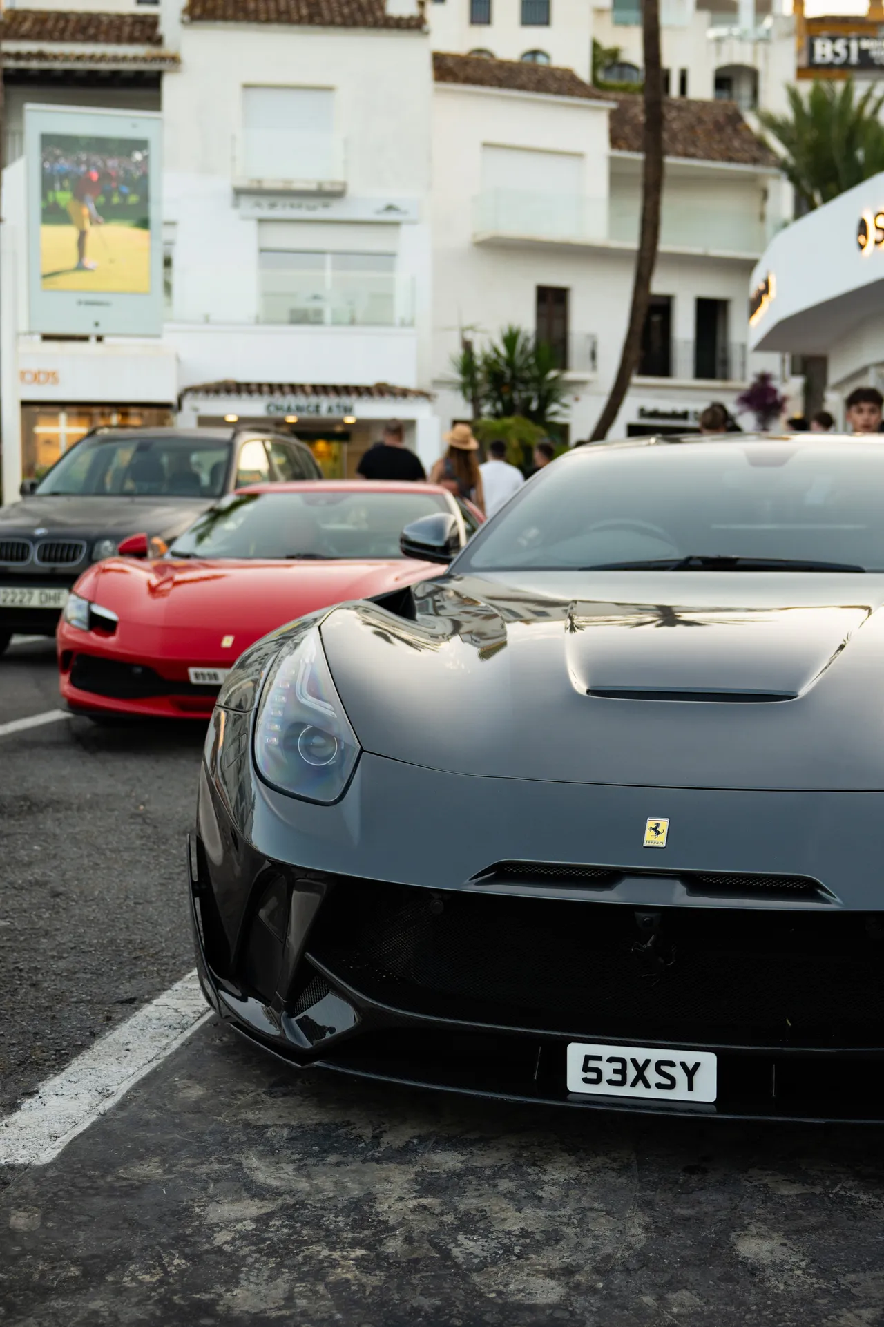 Une Ferrari grise de face au premier plan, une Ferrari rouge garée derrière elle dans une rue commerçante.
