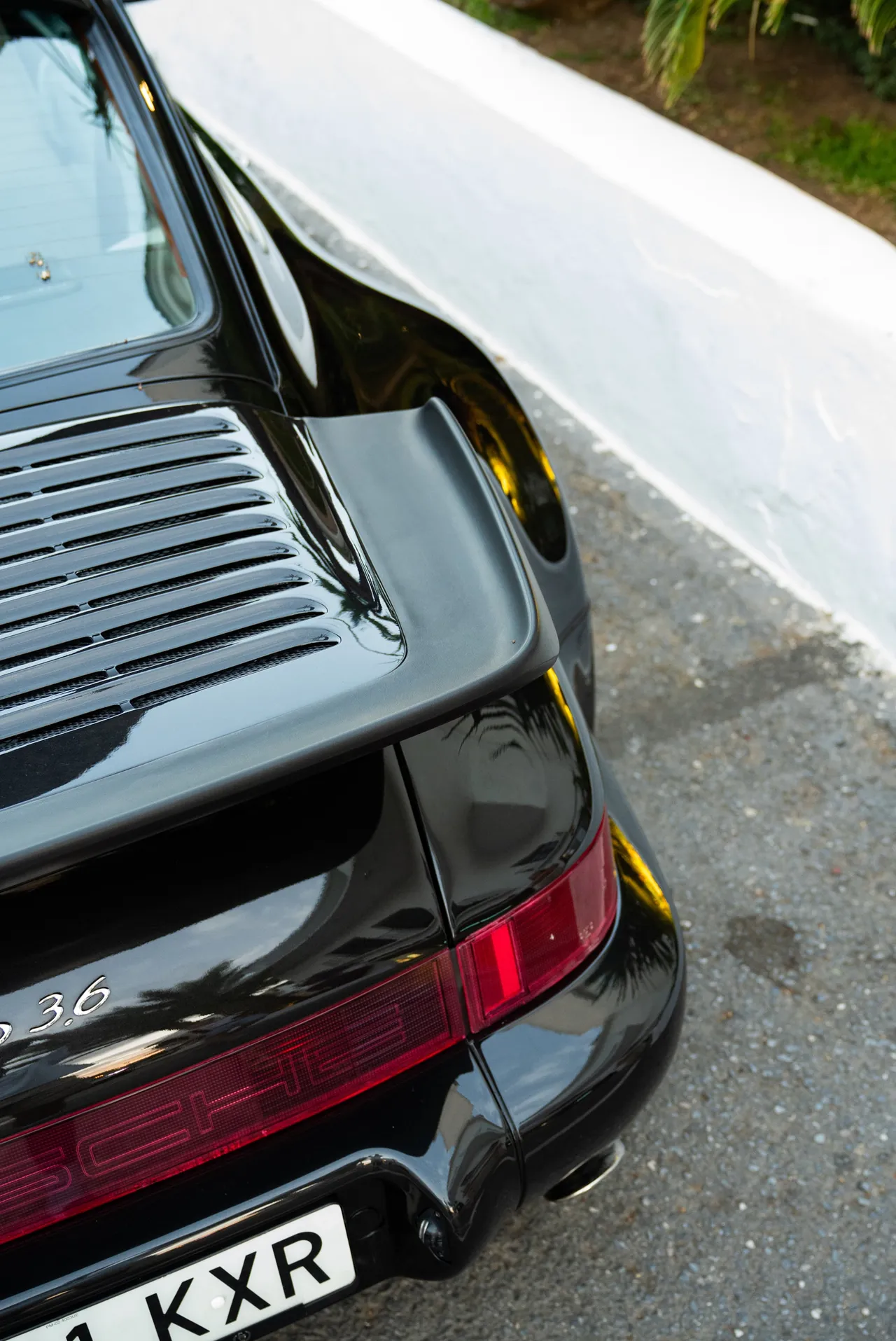 Gros plan sur l’aileron et le feu arrière d’une Porsche 964 Turbo noire, reflets de palmiers sur la carrosserie.